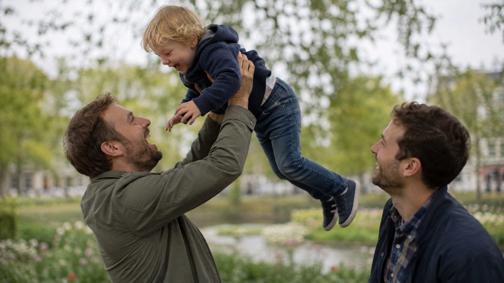 A dad joyfully lifting his laughing child into the air in a park, another dad watching